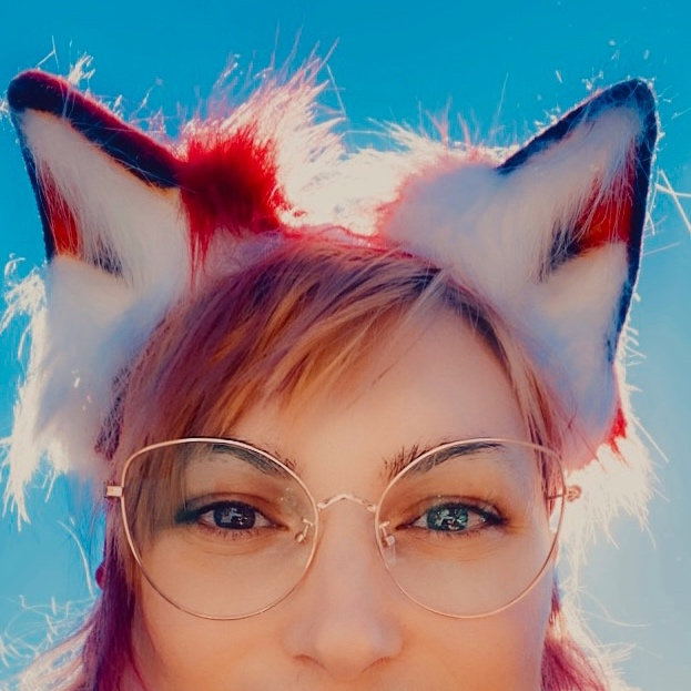 (a photo of my face, cropped at the nose to give the impression of peering over an edge. I am wearing glasses and fox ears in front of a sunny blue sky.)